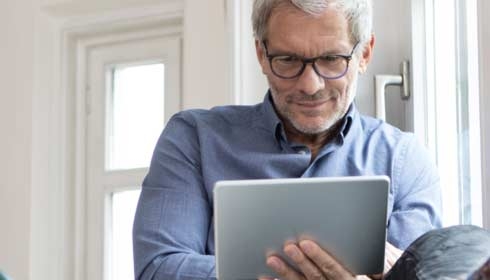 Homme mature à la maison assis à la fenêtre en utilisant une tablette